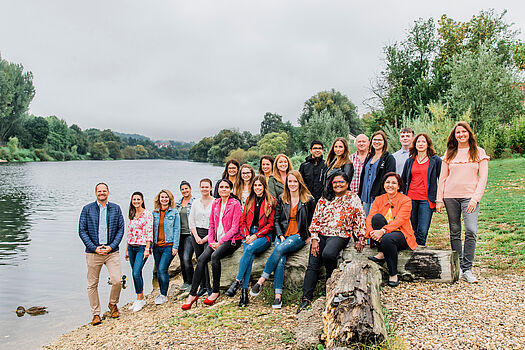 Auf dem Bild sieht man das Team der Geschäftsstelle an einem See, einige sitzen auf einem Baumstamm und andere stehen daneben und dahinter.